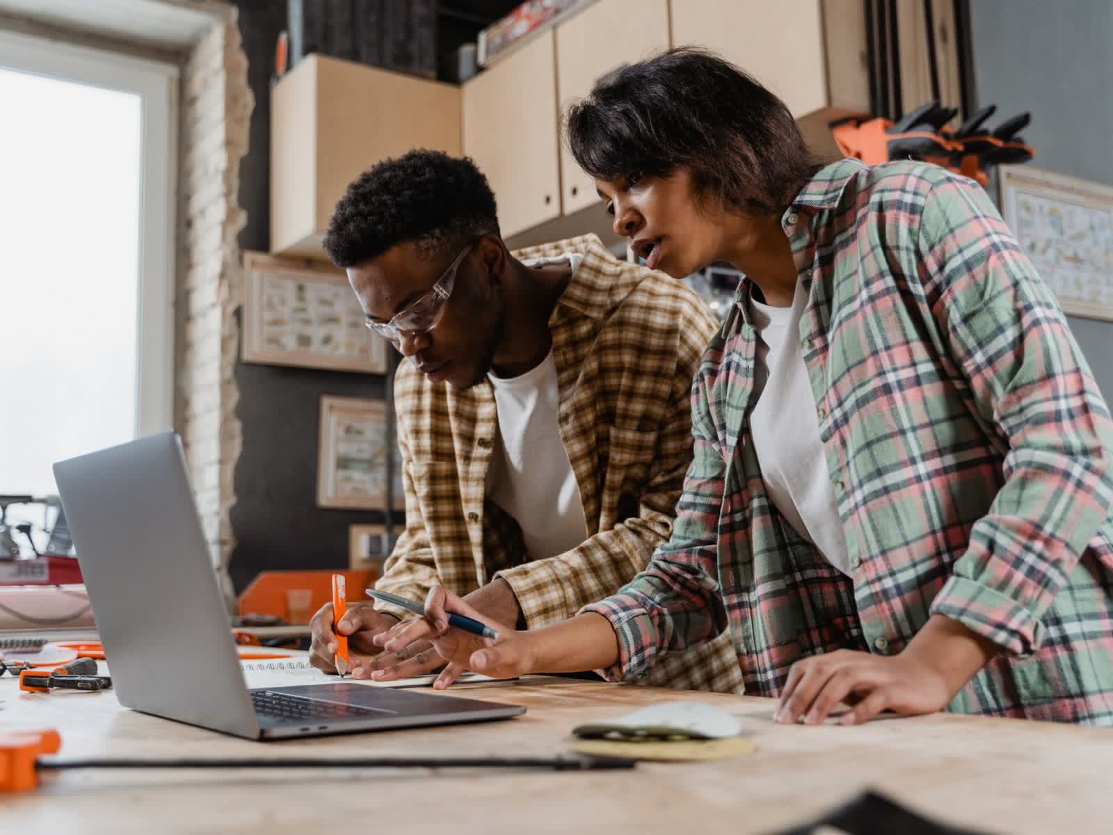 Two people leaning over a workbench in a workshop: they take notes next to a laptop, among tools and materials